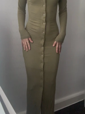 Beige ribbad långklänning med knappar - Säljer en militär grön ribbad långklänning med lång ärm och snygg knappdetalj hela vägen framtill. Klänningen har en tight passform som framhäver figuren och är tillverkad i ett mjukt, stretchigt material. Perfekt för dig som gillar stilrena och enkla plagg med en twist.