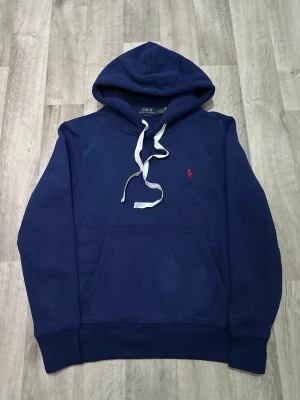 Polo Ralph Lauren mörkblå hoodie  - En mörkblå hoodie från Polo Ralph Lauren. Tröjan har en klassisk passform med huva och vita dragsnören, en känguruficka framtill samt ribbade muddar i ärmslut och nederkant. På vänster bröst sitter den ikoniska broderade Polo-loggan i rött. Tillverkad i mjukt sweatshirtyg