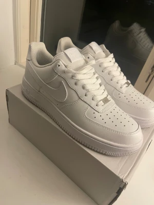 Nike Air Force 1 - Nike Air Force 1 sneakers i helvitt med klassisk låg siluett. Skorna har snörning, perforerad tå och rund tå. Tillverkade i skinn med vit sula och diskret Nike-logga på sidan och hälen. Perfekt för dig som gillar cleana och tidlösa sneakers.