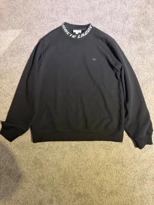 Svart sweatshirt från Lacoste med logga - Svart sweatshirt från Lacoste i klassisk passform. Tröjan har rund hals med vit Lacoste-logga runt kragen och diskret krokodillogo på bröstet. 