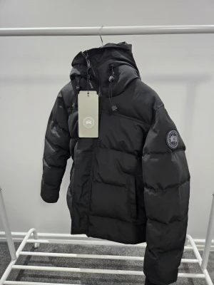 Svart pufferjacka från Canada Goose - Svart pufferjacka från Canada Goose med huva och justerbara snören. Jackan har broderad logotyp på ärmen, dragkedja framtill och quiltad design. Perfekt för kalla dagar och har en clean, modern look som funkar till streetwear.