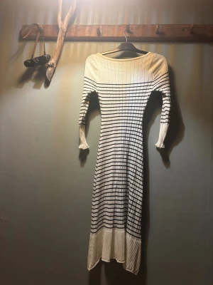 Randig långklänning med v-ringning - Säljer en snygg långklänning i vitt med svarta horisontella ränder. Klänningen har v-ringad hals, långärmade ärmar med volangdetalj vid ärmslut och ribbad struktur. Materialet är stickat och känns mjukt mot huden. Perfekt för dig som gillar klassisk stil med en twist. Formar verkligen sig efter kroppen 💕funkar nog även till xs. 