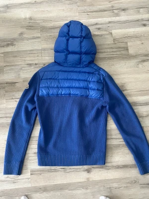 Blå hoodie från Moncler med huva - Säljer en blå hoodie från Moncler med stickad kropp och quiltad, glansig huva och axelparti. Tröjan har långärmade ribbade muddar och en half zip framtill. Moncler-logga på ärmen. Perfekt för dig som gillar exklusiva streetwear-vibbar. Sjukt unik ingen annan som har denna modellen, Äkta såklart. Moncler storlek L vilket motsvarar M/S. Är 180 väger 76 o den sitter perfekt 