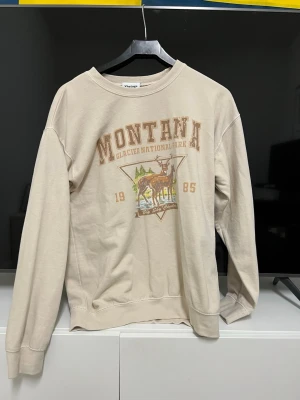 Beige Montana sweatshirt med tryck - Säljer en beige sweatshirt från Vintage med coolt Montana Glacier National Park-tryck och motiv av hjort och natur. Tröjan har rund hals och långa ärmar, perfekt för chill dagar. Trycket har bruna och gröna detaljer som ger en retro känsla.