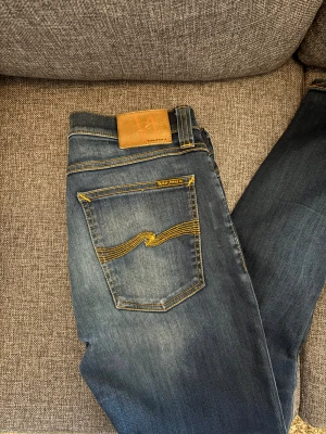 Blå Jeans Nudie Jeans 31/34 - Säljer ett par blå skinny jeans från Nudie Jeans med klassisk femficksdesign och snygga kontrastsömmar i gult. Jeansen har medelhög midja och är tillverkade i mjukt denim med slitningar framtill och bak. Perfekt passform och coola detaljer. Storlek w31 L34 