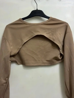 Beige croppad bolerotopp med öppen rygg - Snygg beige bolerotopp med lång ärm och croppad passform. Toppen har en cool öppen ryggdetalj som ger en trendig vibe. Tillverkad i mjukt bomullsmaterial som känns skönt mot huden. Perfekt att styla över linnen eller klänningar för en extra touch.