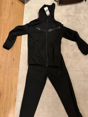 Svart Nike träningsset med huva - Säljer ett svart träningsset från Nike med matchande hoodie och byxor. Hoodien har dragkedja, huva och snygga svarta detaljer över bröstet. Byxorna har dragsko i midjan och smal passform. Perfekt för dig som gillar sportig och stilren look.