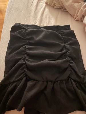 Svart kortkjol med volang - Säljer en svart kortkjol med snygga rynkade detaljer och volang längst ner. Kjolen har en tight passform och är gjord i ett mjukt tyg som känns bekvämt. 