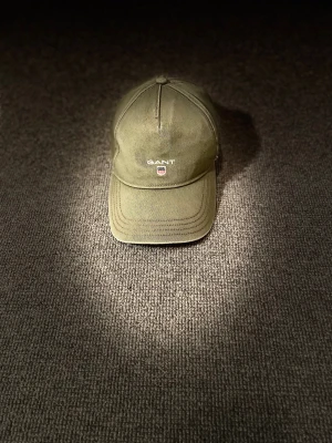 Olivgrön GANT keps med broderad logga - Snygg olivgrön keps från GANT med broderad vit logga och sköld framtill. Klassisk böjd skärm och justerbar rem baktill för perfekt passform. Tillverkad i slitstark bomull, perfekt för dig som vill ha en clean och stilren look. Man kan också matcha den med väldigt många outfits! Skriv till mig om du undrar om något! [Ganska sliten]