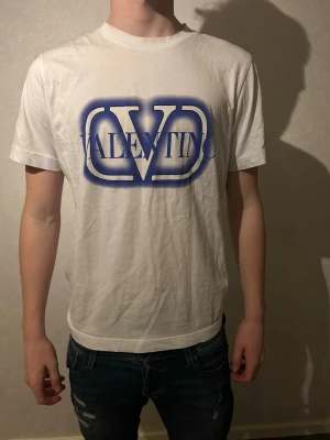 Vit Valentino t-shirt med blå logga - Vit t-shirt från Valentino med stor blå logga framtill. Klassisk rund halsringning och korta ärmar. Tillverkad i mjuk bomull för en skön känsla. Perfekt för dig som vill ha en clean men ändå statement look. Har kvar lapp och knappt använd