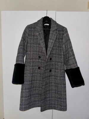 Rutig kappa från Noella med pälsdetalj - Säljer en snygg rutig kappa från Noella i storlek S. Den har svart fuskpäls vid ärmsluten, dubbelknäppning och klassisk krage. Färgen är grå och svart med ett stilrent rutmönster. Perfekt för dig som vill ha en statement jacka med coola detaljer.