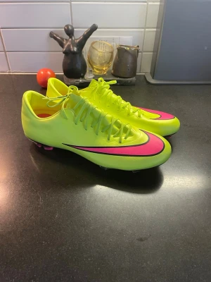 Nike Mercurial Vapor - Säljer ett par grymma Nike fotbollsskor i neon-gult med rosa detaljer och svart Nike-logga. Skorna har snörning, platt sula med dobbar och är tillverkade i syntetmaterial. Perfekt för dig som vill sticka ut på planen och ha maxad kontroll på bollen.
