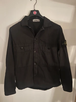 Svart overshirt från Stone Island - Svart overshirt från Stone Island med klassisk krage, två bröstfickor med knappar och ikonisk patch på vänster ärm. Jackan har knappstängning framtill och är tillverkad i bomull. Perfekt för dig som gillar clean och stilren streetwear.