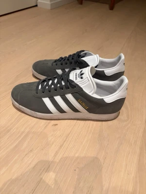 Adidas Gazelle sneakers grå/vita - Säljer ett par Adidas Gazelle sneakers i grå mocka med vita detaljer och klassiska tre ränder på sidorna. Skorna har snörning, vit plös med Adidas-logga och rund tå. Insidan är vit och yttersulan är platt. Använda 2 gånger storlek 42