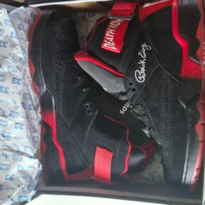 Patrick Ewing Death Row sneakers svart/röd - Limited edition Patrick Ewing Death Row Records sneakers i svart och rött med högt skaft, snörning och bred rem med text. Snygga detaljer som Death Row-logga och Patrick Ewings signatur. Materialet är mocka och syntet, med platt sula och rund tå.