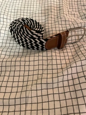 Svartvit flätad bälte med bruna detaljer - Snyggt flätat bälte i svart och vitt med bruna läderdetaljer och silvrigt spänne. Bältet har ett coolt zigzag-mönster och passar perfekt till jeans eller chinos. Den bruna läderdelen har vit söm som ger en extra touch.