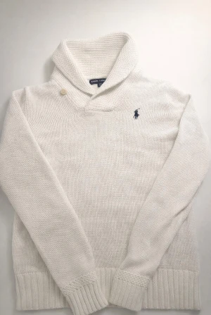 Vit stickad tröja från Ralph Lauren - Vit stickad tröja från Ralph Lauren med grovstickad krage och en knappdetalj vid halsen. Tröjan har lång ärm och ribbade muddar samt en broderad marinblå logga på bröstet. Perfekt för dig som gillar klassisk och stilren design.