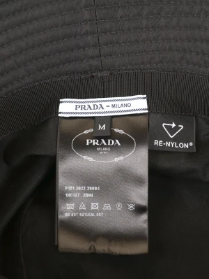 Svart bucket hat från Prada - Snygg svart bucket hat från Prada med ikonisk metalltriangel-logga framtill. Hatten är tillverkad i glansig re-nylon som ger en modern vibe och har diskreta sömmar runt brättet. Perfekt accessoar för att lyfta din streetstyle. Har aldrig använts! 