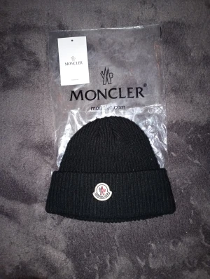 Moncler mössa - Säljer en svart ribbstickad mössa från Moncler med klassisk logotyp framtill. Mössan är tillverkad i mjuk ull och har en bred uppvikt kant för extra stilpoäng. Perfekt för dig som vill ha en clean och snygg look under kyliga dagar. Syd på toppen men syns inte, se bild 3. Gratis marmor armband vid köp!