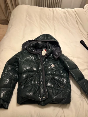 Mörkgrön pufferjacka från Moncler - Snygg mörkgrön pufferjacka från Moncler med glansig finish och huva. Jackan har quiltad design, knappar framtill och praktiska fickor med dragkedja. Perfekt för kalla dagar och har en ikonisk Moncler-logga på bröstet.