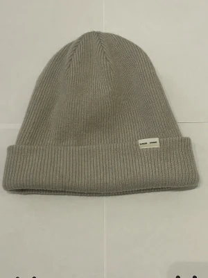 Ljus grå  stickad mössa från Samsøe Samsøe - Snygg ljust grå  mössa från Samsøe Samsøe i ribbstickad design. Mössan har en uppvikt kant med en liten vit etikett med loggan framtill. Tillverkad i mjukt material som känns skönt mot huden och passar perfekt för kyliga dagar.