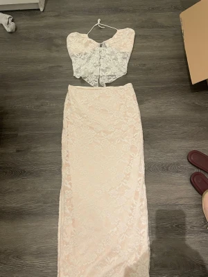 Vit spetsig omlott korsett - Säljer en vit omlott korsett med blommigt spetsmönster och smala axelband. Toppen har en croppad passform och öppen rygg, vilket ger en snygg och modern look. Perfekt att matcha med kjol eller jeans för en trendig outfit.