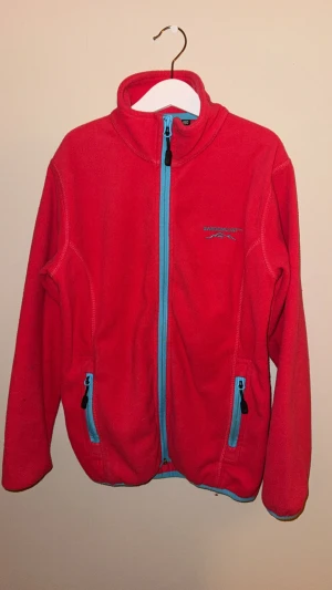 Röd fleecejacka från Swedemount - Röd fleecejacka från Swedemount med blå dragkedjor och detaljer. Jackan har hög krage, två fickor med dragkedja och är tillverkad i 100% polyester. Perfekt för kyliga dagar och enkel att slänga på sig över en hoodie.