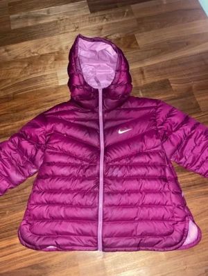 Mörklila pufferjacka från Nike - Säljer en snygg mörklila pufferjacka från Nike med huva och dragkedja. Jackan har ett quiltat mönster och är fodrad i ljusrosa. Perfekt för kalla dagar och har en sportig vibe med Nike-loggan på bröstet.