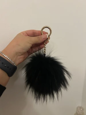 Svart pälsboll från Boliris - Säljer en fluffig svart pälsboll från hollies med guldfärgad kedja och ring. Perfekt som accessoar till väskan eller nyckelknippan. Pälsbollen är rund och har en lyxig känsla tack vare den glansiga gulddetaljen.