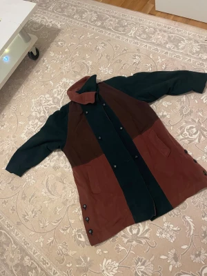 Brun och mörkgrön rock kappa  från Scandinavian Look - Unik kappa  i brun och mörkgrön färgblock från Scandinavian Look, storlek 40. Jackan har dubbelknäppning med svarta knappar, två fickor fram och en mjuk, glansig brun insida. Perfekt för dig som gillar retrovibbar och vill sticka ut med stil.