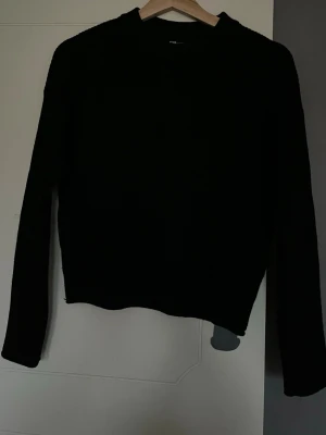 Svart ribbad stickad tröja - Svart stickad tröja med ribbad struktur och lång ärm. Modellen har en klassisk rund hals och är något croppad i passformen. Perfekt för dig som gillar enkla och stilrena plagg med lite edge. Snygg att matcha med jeans eller kjol.