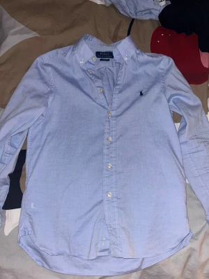 Ljusblå skjorta Polo Ralph Lauren - Snygg ljusblå skjorta från Polo Ralph Lauren i slim fit-modell. Klassisk button-down krage, broderad logga på bröstet och vita knappar framtill. Tillverkad i mjuk bomull som är skön mot huden. Perfekt för dig som gillar stilrena och tidlösa plagg.