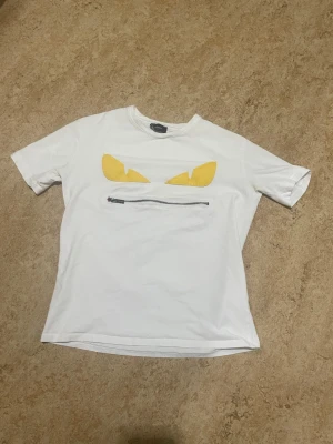 Vit Fendi t-shirt med dragkedja - Säljer en vit t-shirt från Fendi med coola gula applikationer som ser ut som ögon och en silverfärgad dragkedja på bröstet. T-shirten har rund hals och korta ärmar, tillverkad i mjuk bomull. Perfekt för dig som gillar statement-plagg och designer.  #fendi #fenditshirt #designer #designertshirt #lyx #luxury #streetwear #highend #fashion #mode #stil #plick #plicksale #plickfynd #secondhand #vintage #sverige #sweden #ungdomsmode