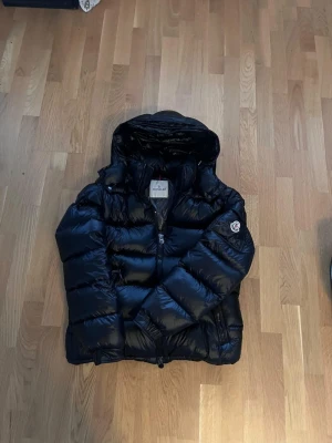 Moncler maya - Säljer en svart pufferjacka från Moncler med glansig finish och huva. Jackan har tvåvägsdragkedja med Moncler-logga, samt logotyp på ärmen. Fylld med dun och har en loose passform, perfekt för kalla dagar. Flera fickor med dragkedja och snygga detaljer. STORLEK 2, mycket bra skick,inga defekter.