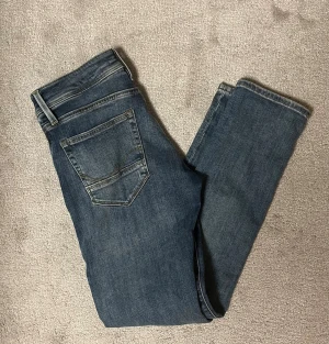 Blå slitna jeans från Jack & Jones - Säljer ett par blå jeans från Jack & Jones med snygga slitningar och lagade hål på benen. Jeansen har klassisk femficksdesign, normal passform och är tillverkade i jeansmaterial med lite stretch. Perfekt för dig som gillar en avslappnad och trendig stil.