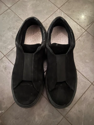 Svarta slip-on sneakers i mocka/ Legend Kane sneakers. - Svarta slip-on sneakers med bred resår över vristen för enkel påtagning. Skorna har en platt sula och är tillverkade i mocka, vilket ger en stilren och clean look. Perfekta för dig som gillar minimalistisk stil och vill ha något som funkar till det mesta.                             skor: Legend Kane sneakers. Ny pris 1500. Skorna är inte Arigatos.