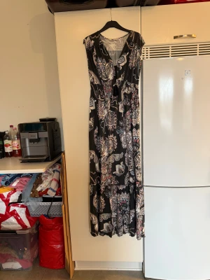 Svart långklänning Odd Molly blommig - Långklänning från Odd Molly med svart botten och stora blommiga mönster i vitt, beige och blått. Klänningen har v-ringning, knytband med tofsar och smock i midjan. Luftigt material och ärmlös design, perfekt för en avslappnad och snygg look. Från märket Odd Molly. Storlek 0 som enligt deras storleksguide motsvarar en XS. Men passar mer en M. Mer använd. Kanske kan vara något blekare än ny. Men fortfarande otroligt fint skick. 