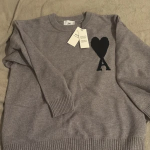 Grå stickad tröja från Ami - Snygg grå stickad tröja från Ami med svart hjärta och bokstaven A broderat på bröstet. Tröjan har rund hals, ribbade muddar och är långärmad. Perfekt för dig som gillar stilrena detaljer och vill ha något som sticker ut lite.