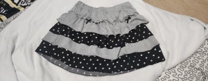 Grå och svart volangkjol med prickar stl. S/M - Supersöt kort volangkjol i grått och svart med vita prickar. Kjolen har flera lager med volanger och ett elastiskt midjeband för skön passform. Perfekt för dig som gillar en lekfull och trendig stil. Små rosetter framtill ger extra gullig detalj.
