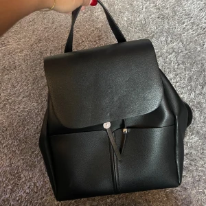 Svart ryggsäck i skinnimitation från Zara - Stilren svart ryggsäck från Zara i skinnimitation med slät yta och minimalistisk design. Väskan har lock, snörning och justerbara axelremmar. Perfekt för dig som vill ha en clean och modern look till vardagen.