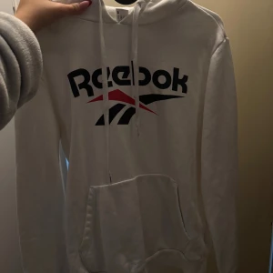Vit hoodie från Reebok med logga - Vit hoodie från Reebok med stor svart och röd logga framtill. Klassisk känguruficka, huva med snörning och långa ärmar. Perfekt för en sportig och chill stil.