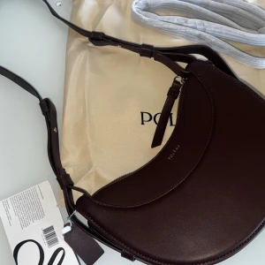 Polène brun axelväska i skinn - För föer bilder och info skicka ett meddelande. Snygg halvmåneformad axelväska  i mörkbrunt skinn. Väskan har justerbar axelrem (ett längre axelrem följer med, guldfärgade detaljer och dragkedja upptill. Perfekt för dig som gillar stilrena accessoarer med exklusiv känsla.