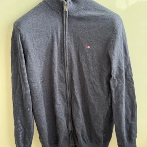 Grå kofta med dragkedja från Bison - Mörkgrå kofta från Bison med hel dragkedja och liten broderad logga på bröstet. Tröjan har ribbad krage, muddar och är tillverkad i mjukt material som passar perfekt till lager-på-lager. Enkel och stilren design för en clean look.