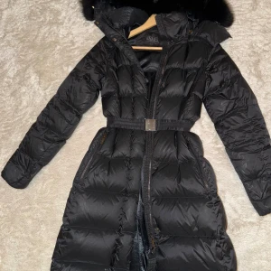 Svart lång dunjacka från Áhkká  - Snygg svart lång dunjacka från Áhkká med bälte i midjan och stor huva med päls. Jackan har dragkedja framtill och två fickor med dragkedja. 