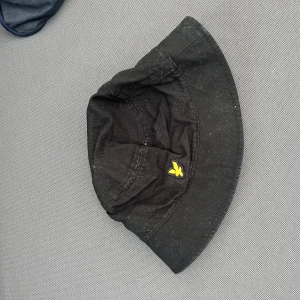 Svart bucket hat från Lyle & Scott - Svart bucket hat från Lyle & Scott med klassisk gul logga framtill. Hatten har bred brätte och är tillverkad i bomull, perfekt för en avslappnad streetstyle-look.