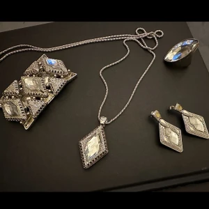 Set med silverfärgade smycken med kristaller - Säljer ett komplett set med halsband, armband, ring och örhängen i silverfärg med stora, slipade kristaller. Alla delar har en cool geometrisk design med rektangulära och diamantformade detaljer, perfekt för dig som gillar statement-smycken.