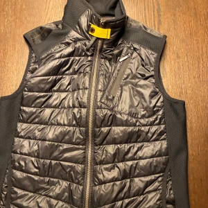 Svart dunväst från Parajumpers - Svart dunväst från Parajumpers med quiltad design och gul detalj vid kragen. Västen har dragkedja framtill, bröstficka med dragkedja och sportig känsla. Perfekt för lager-på-lager och har en stilren look med diskreta sömmar.