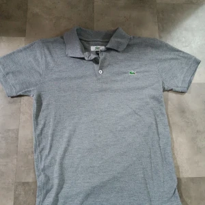 Grå pikétröja från Lacoste - Snygg grå pikétröja från Lacoste med klassisk krage och korta ärmar. Tröjan har den ikoniska gröna krokodilloggan på bröstet och stängs med två knappar framtill. Perfekt för en clean och sportig stil.