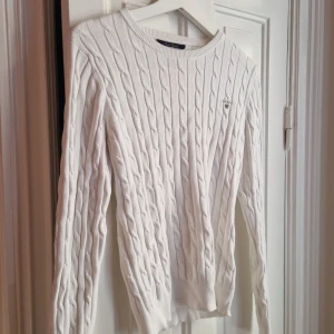 Vit kabelstickad tröja från Gant - Klassisk vit kabelstickad tröja från Gant i stretchig bomull. Klassisk rund halsringning, diskret Gant-logo på bröstet och ribbade muddar. Perfekt för dig som gillar en clean och tidlös stil med snygga detaljer.