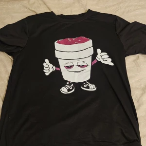 Svart t-shirt Y2K - Svart t-shirt med stor grafisk print av en tecknad kopp med sneakers på framsidan och texten 'THAT'S A AWFUL LOT OF COUGH SYRUP' i lila och vitt på baksidan. T-shirten har rund hals och korta ärmar. Materialet känns som bomull och passformen är normal.